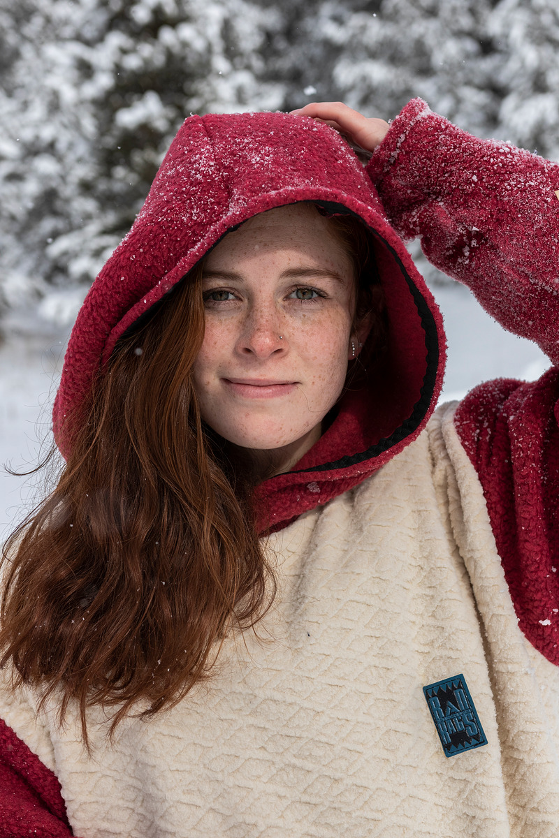Freckled model in a cream sherpa hoodie with a maroon hood, snow falling