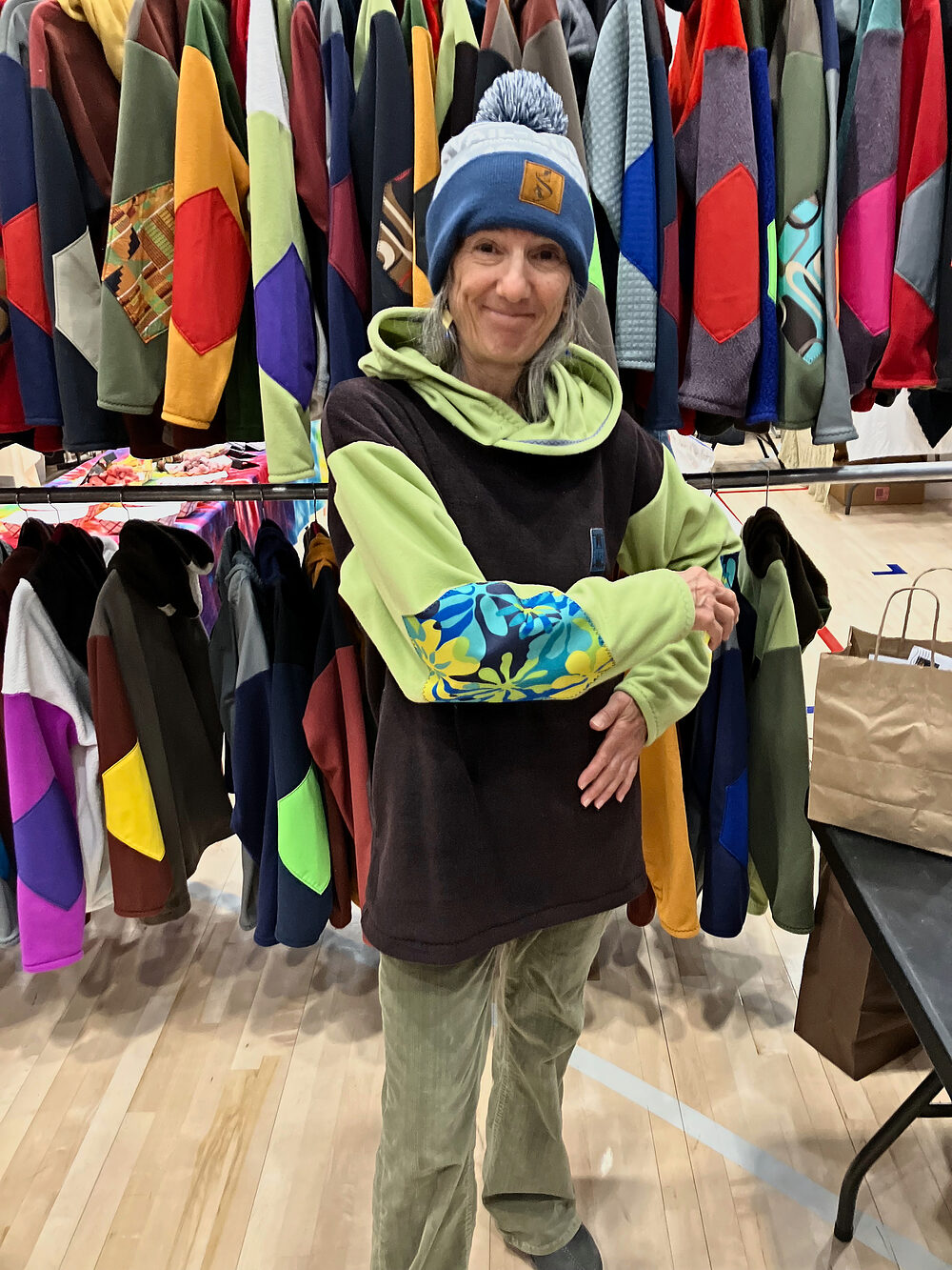 Woman in a black and green Rad Rags hoodie with a floral sleeve and a Rad Rags beanie at an indoor pop-up, racks of fleece behind her