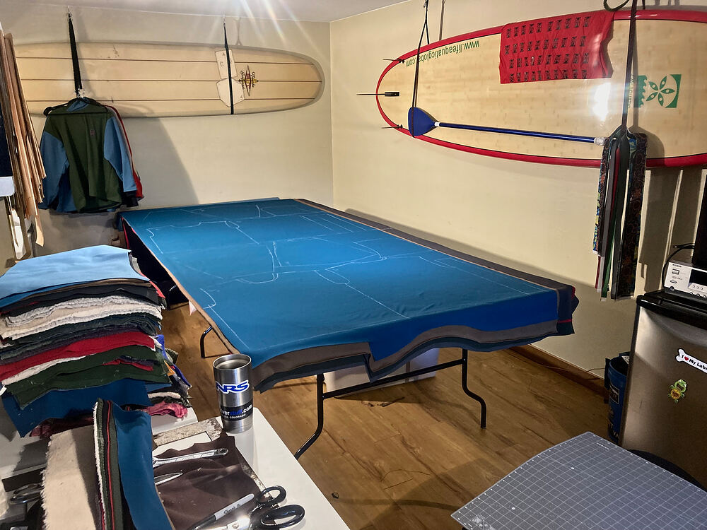 The workshop: a blue cutting table and a stack of fleece, with surfboards hung on the wall above