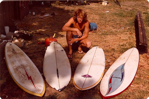 Vintage photo from Sunset Beach, Oahu, in 1979: a shirtless young surfer with curly hair crouched behind four surfboards