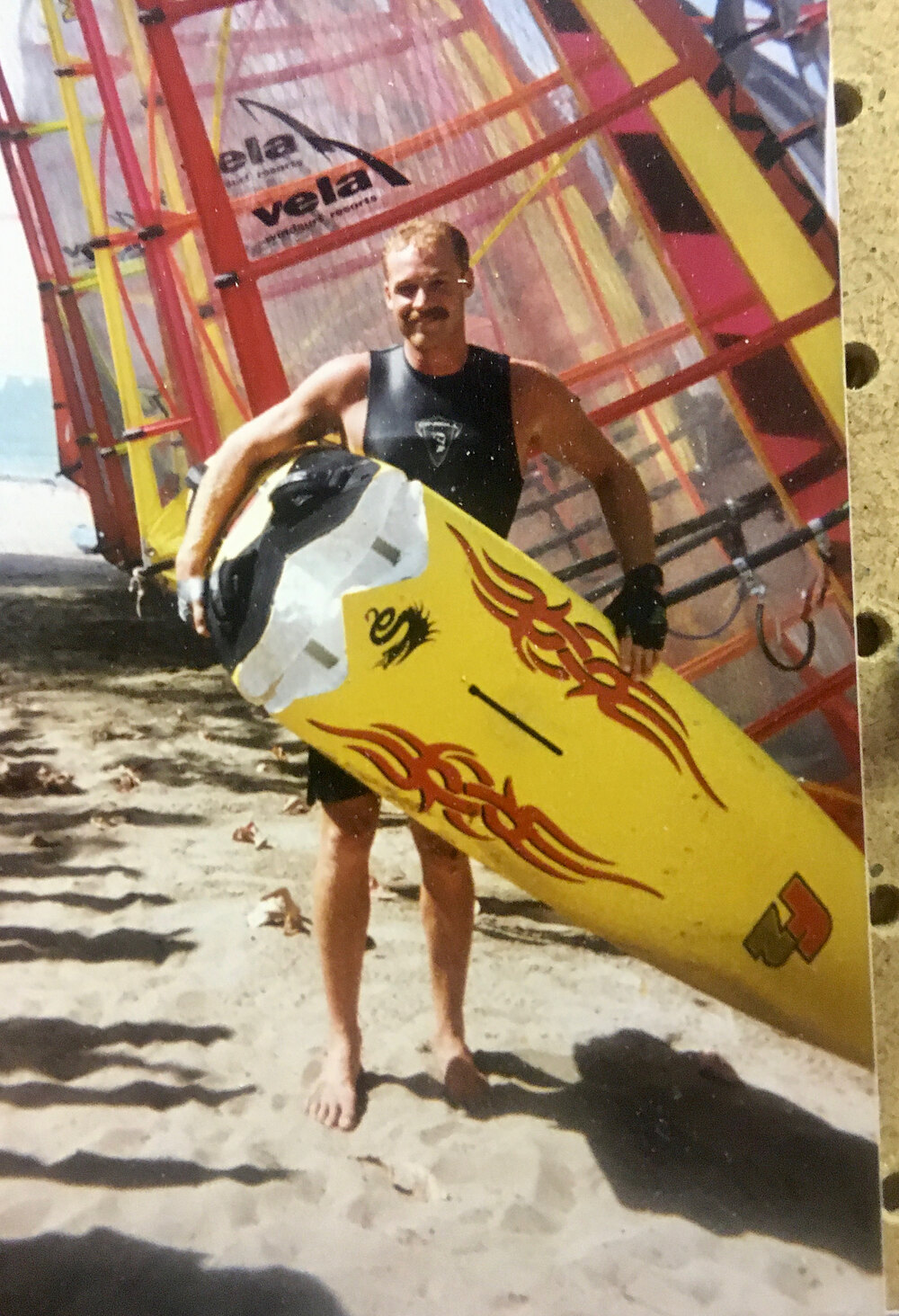 Old photo of a man holding a yellow windsurf board on a beach in the Dominican Republic, with sails behind him