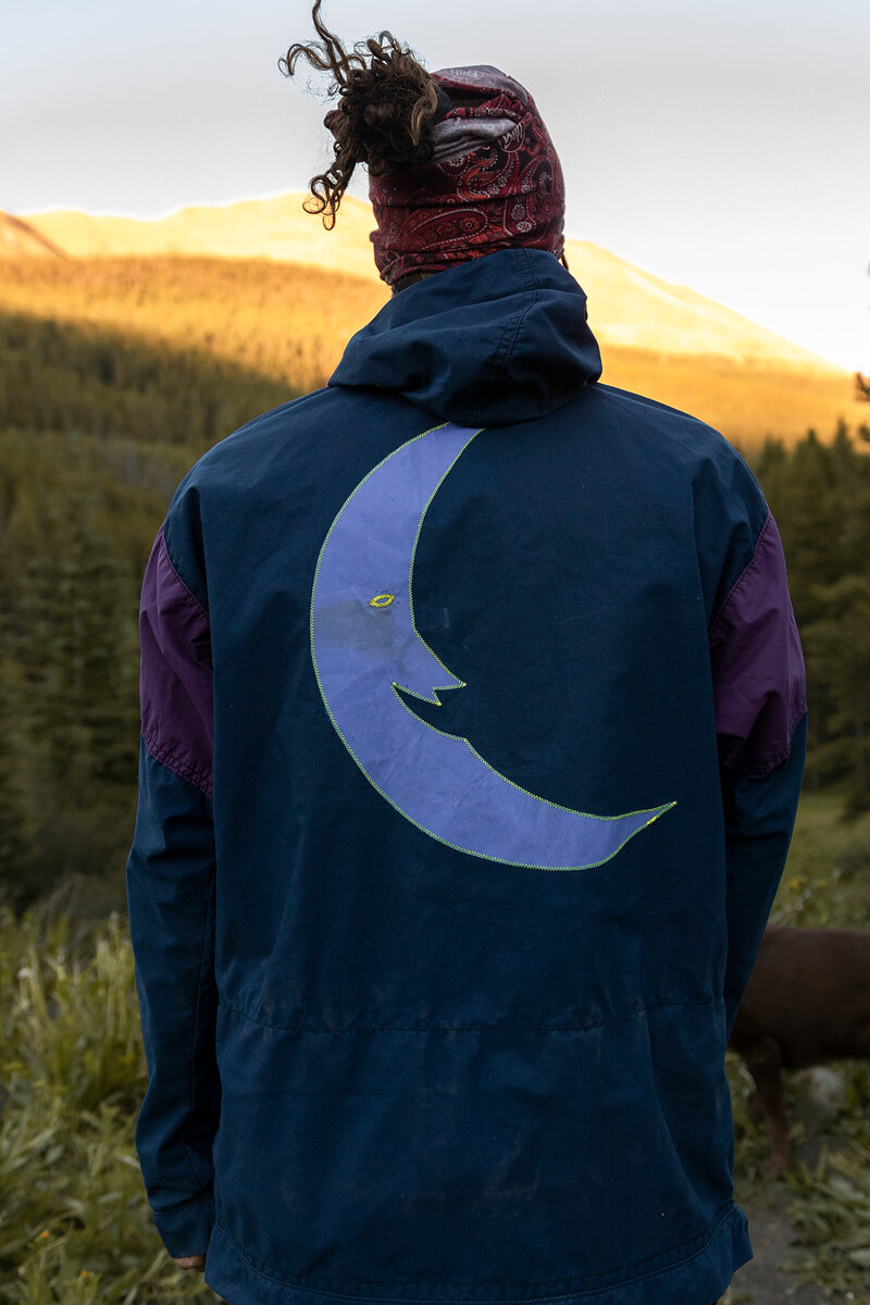 Back of a navy hooded jacket with a big lavender crescent moon, mountains in golden light