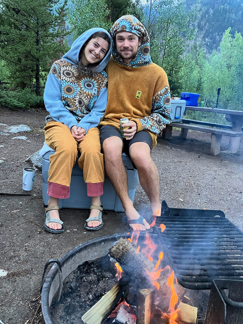 Two friends in matching sunflower-print Rad Rags hoodies by a campfire