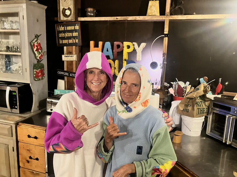 Two women in Rad Rags hoodies with the hoods up, throwing shakas in a kitchen