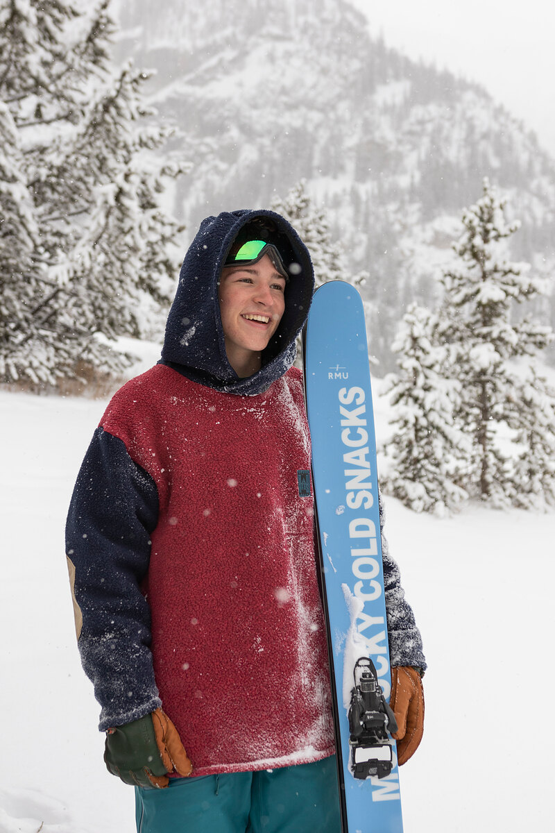 Smiling model in a maroon and navy color-block hoodie holding a ski
