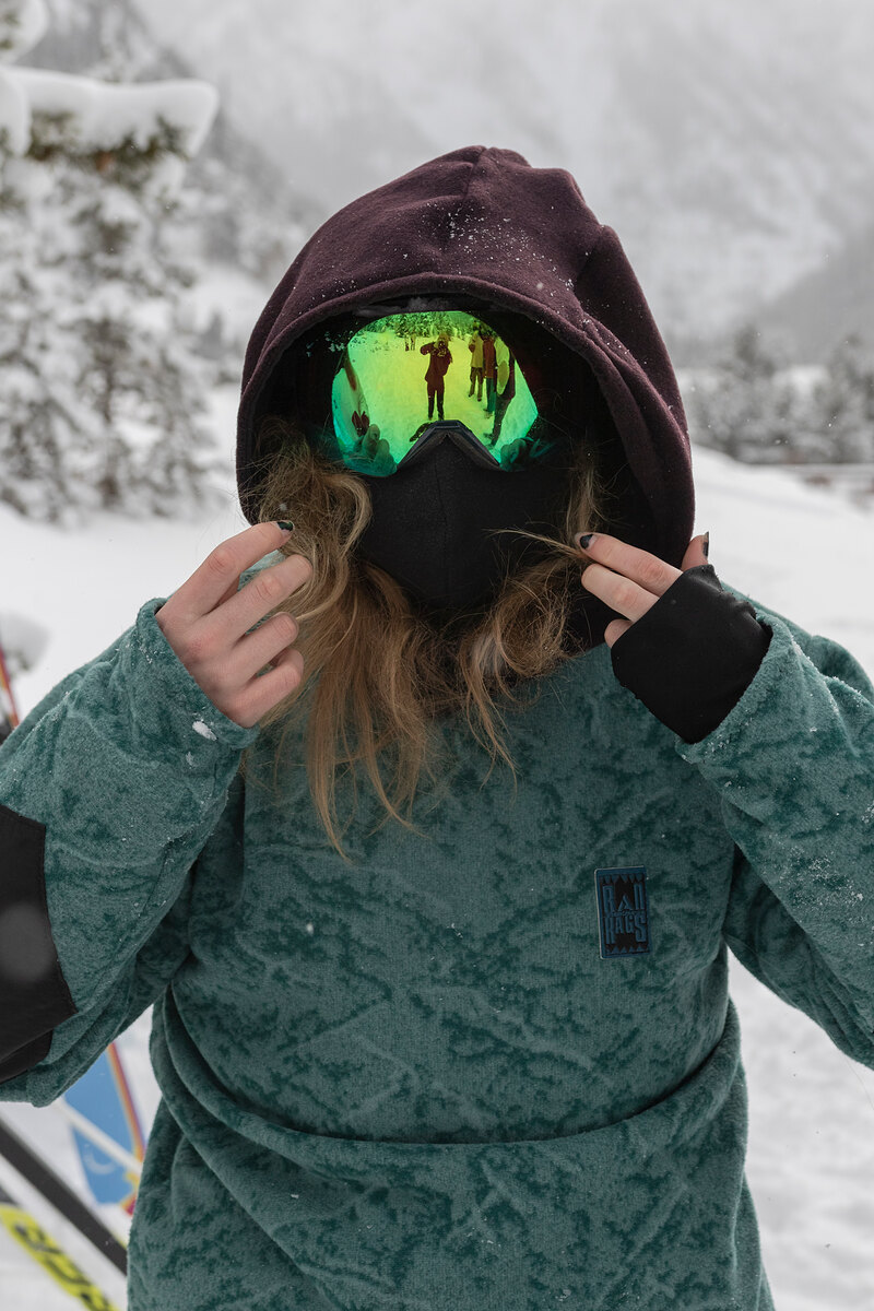 Model in a green print fleece pulling up the hood, mirrored goggles on