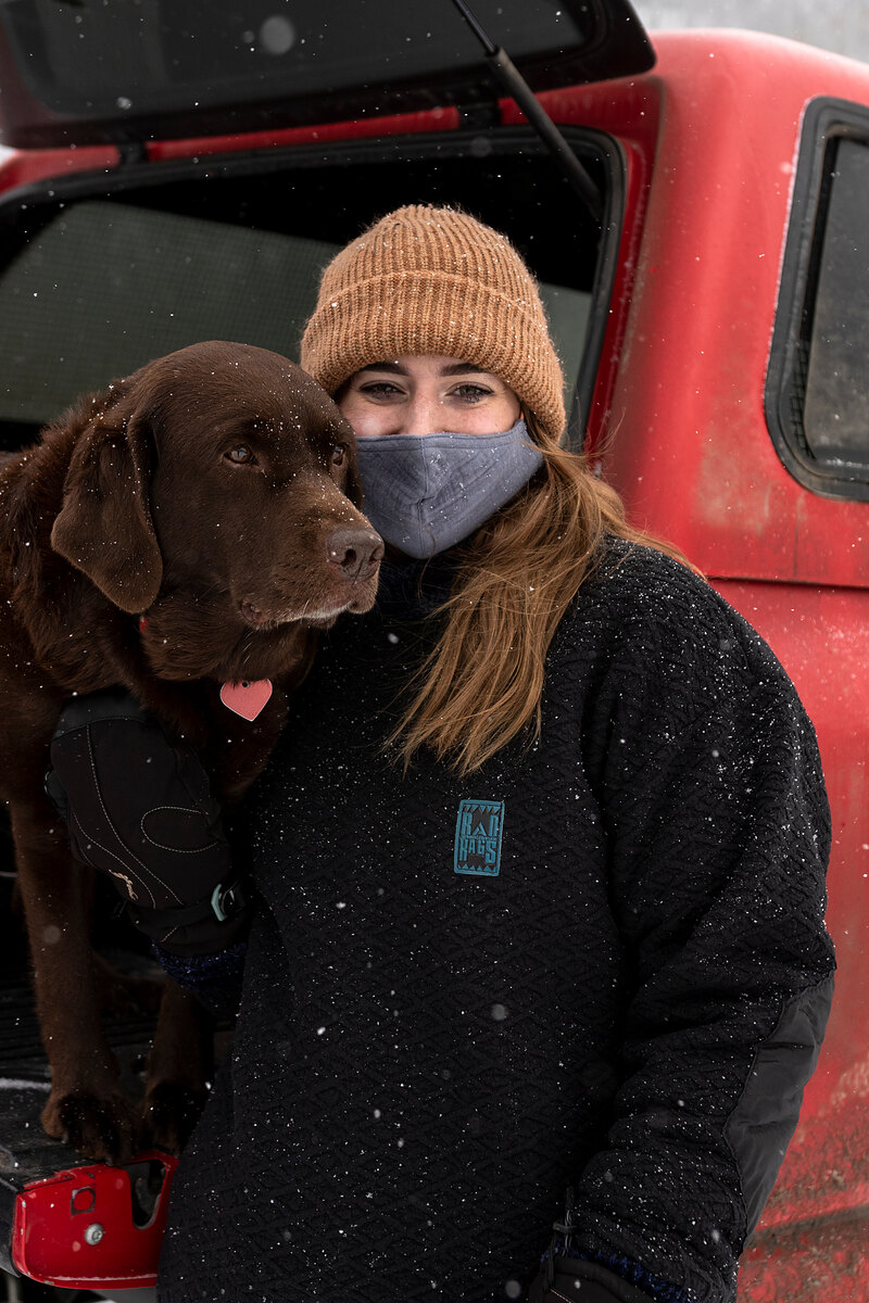 Model in a black fleece hugging a chocolate lab beside a red truck
