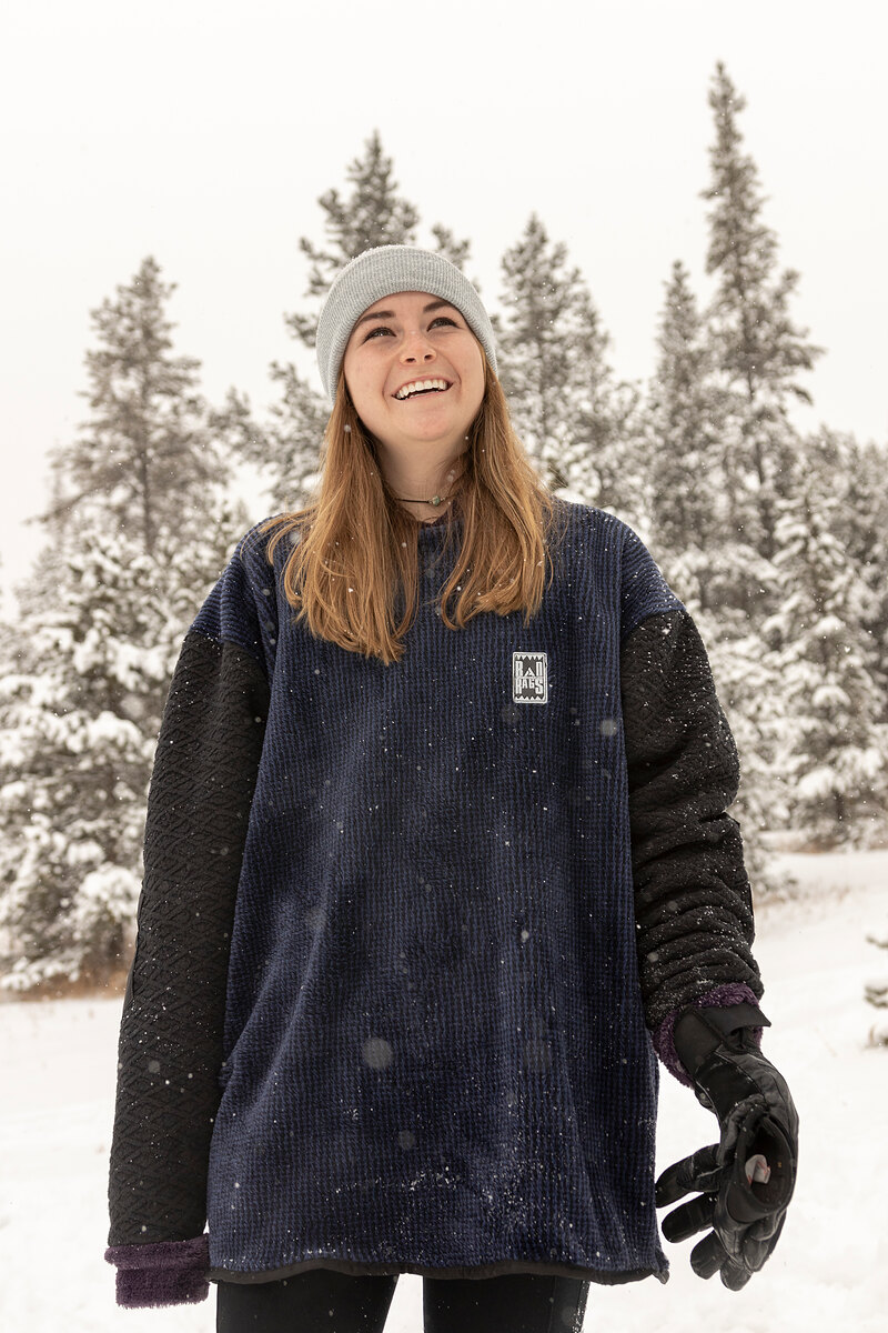 Smiling model in a navy fleece with black sleeves among snowy trees