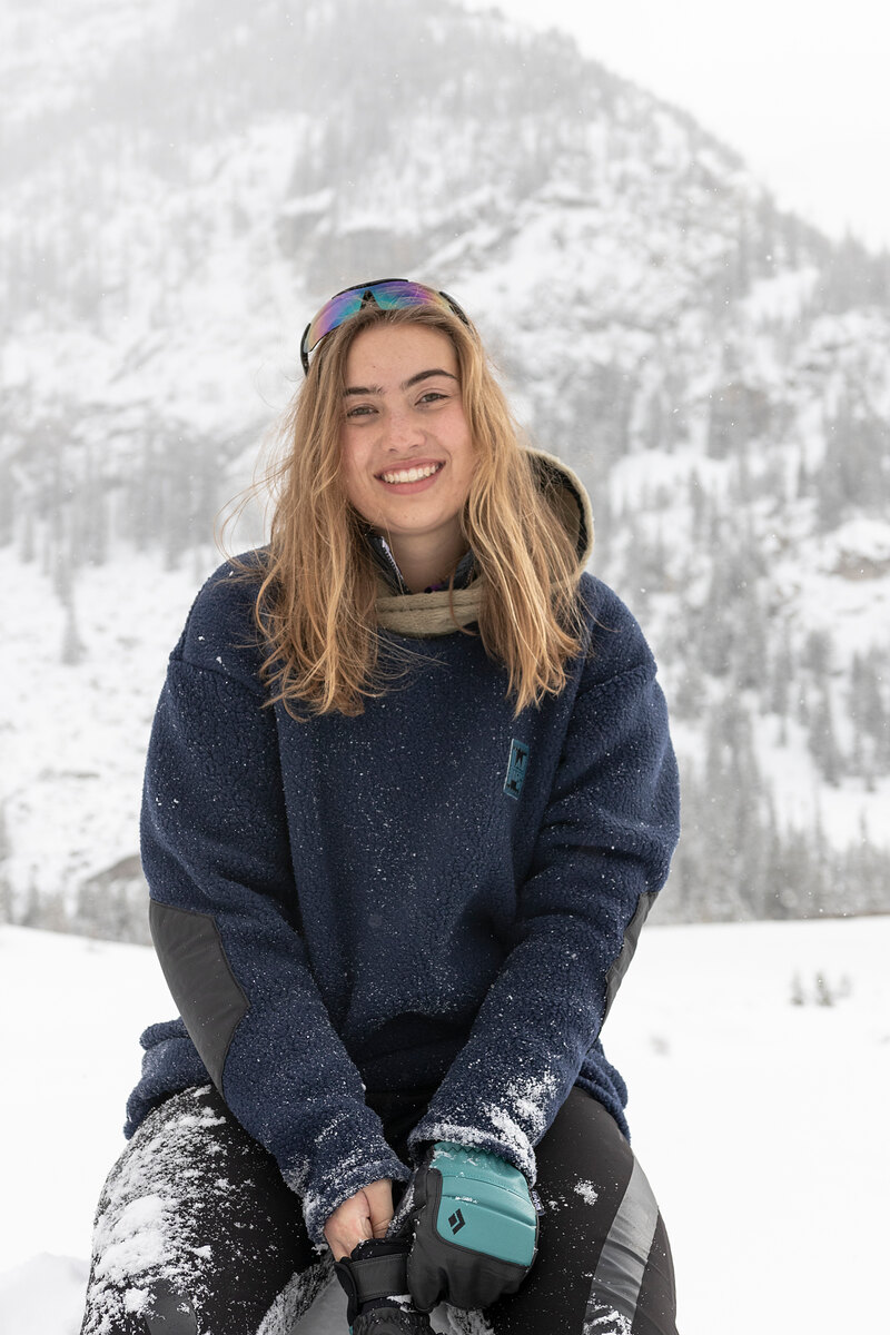 Model in a navy fleece sitting in the snow, goggles pushed up